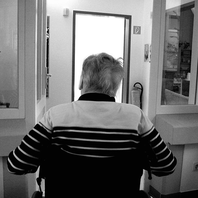 Older man in a wheelchair indoors, viewed from behind.